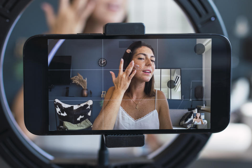 Woman applying makeup is visible on a phone screen set up on a ring light. Background includes a clock and wall decor