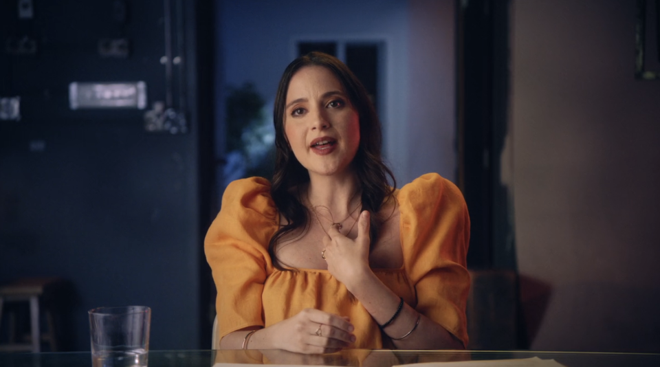 Alexa in orange dress sitting at table with hand on chest, expressive face, indoors