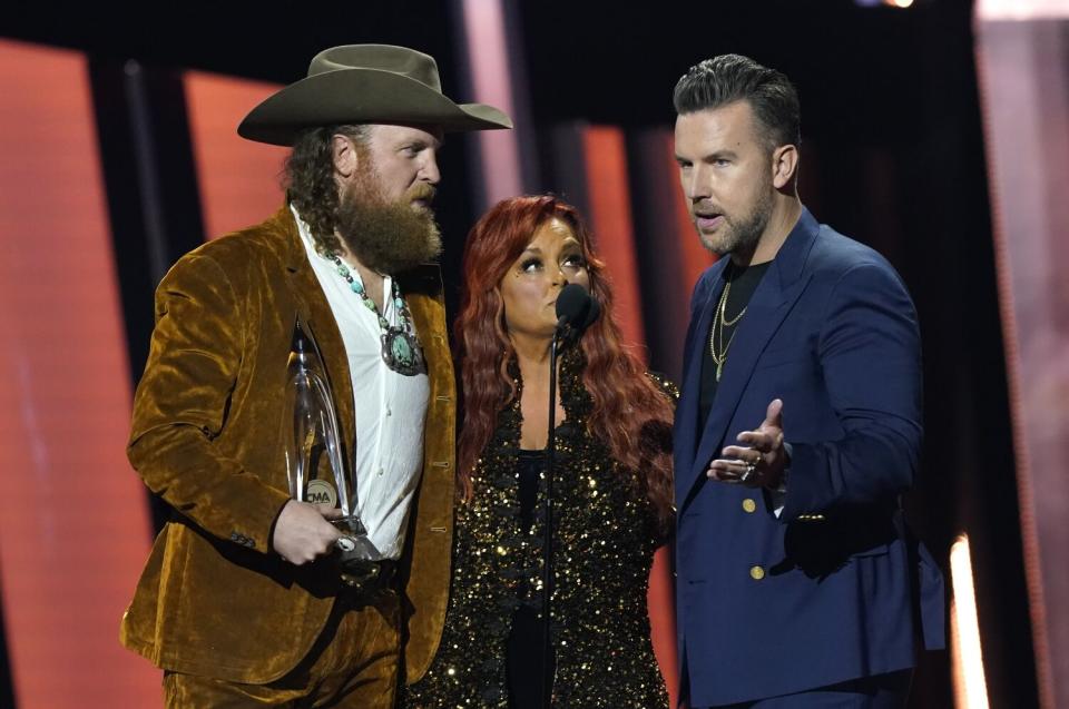 A woman with red hair stands between two men onstage