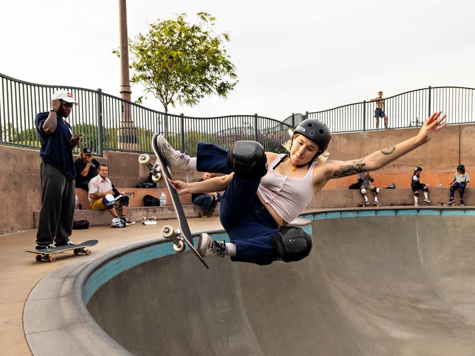 A woman skates above a bowl on the left side with people watching behind her.