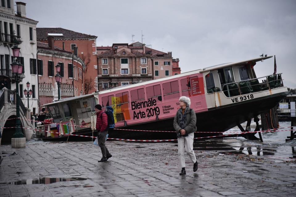 TOPSHOT - People walk past a stranded taxi boat on Riva degli Schiavoni, after it was washed away during an exceptional overnight "Alta Acqua" high tide water level, early on November 13, 2019 in Venice. - Powerful rainstorms hit Italy on November 12, with the worst affected areas in the south and Venice, where there was widespread flooding. Within a cyclone that threatens the country, exceptional high water were rising in Venice, with the sirocco winds blowing northwards from the Adriatic sea against the lagoons outlets and preventing the water from flowing back into the sea. At 22:40pm the tide reached 183 cm, the second measure in history after the 198 cm of the 1966 flood. (Photo by Marco Bertorello / AFP) (Photo by MARCO BERTORELLO/AFP via Getty Images)