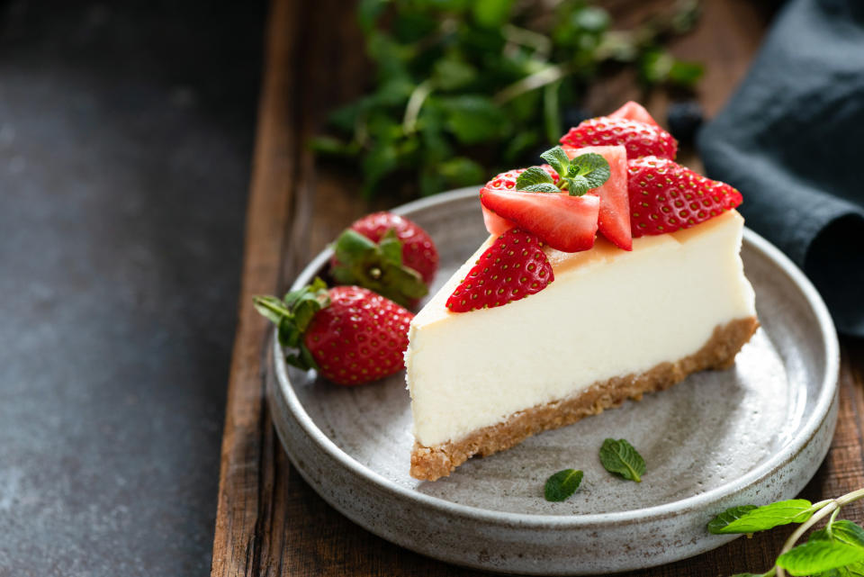 A slice of cheesecake on a plate topped with sliced strawberries and a sprig of mint, two strawberries and mint leaves on the side, all on a wooden plant with mint and a cloth napkin in the background