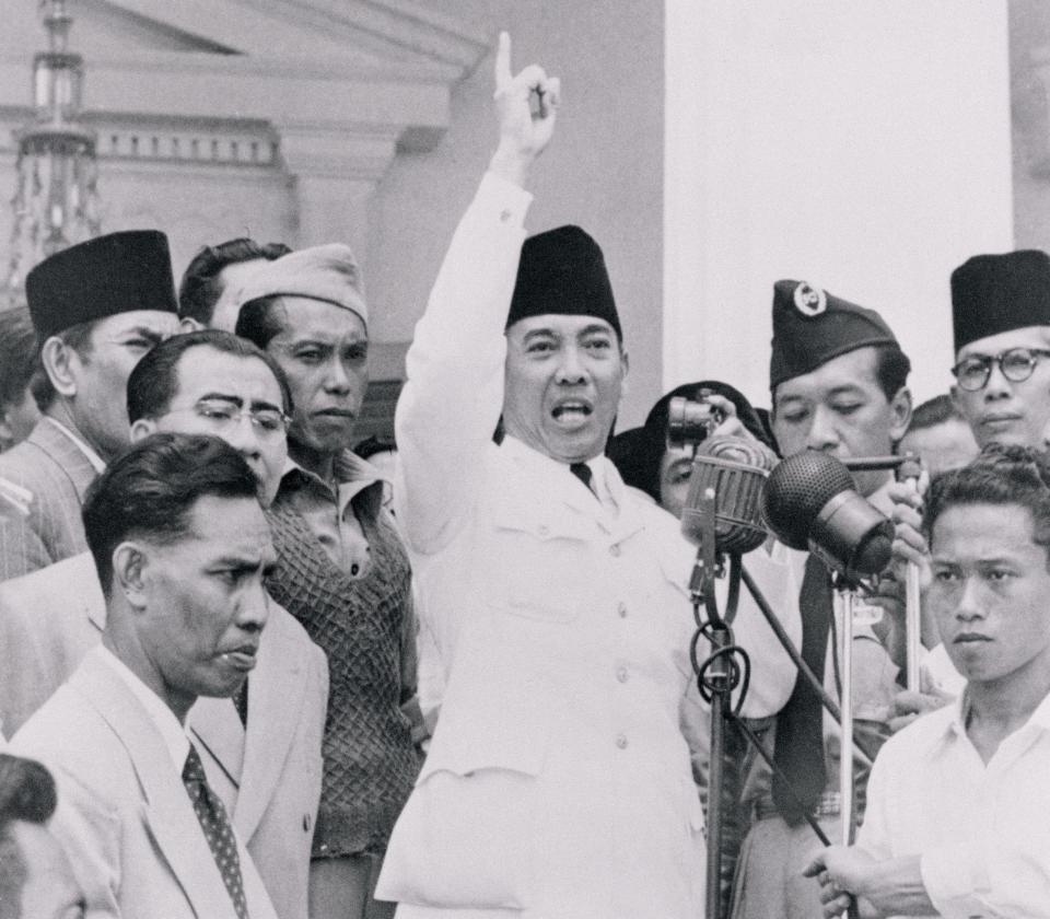 A photo of a man dressed in a white suit with a hand raised in a pointed finger, speaking into many microphones.