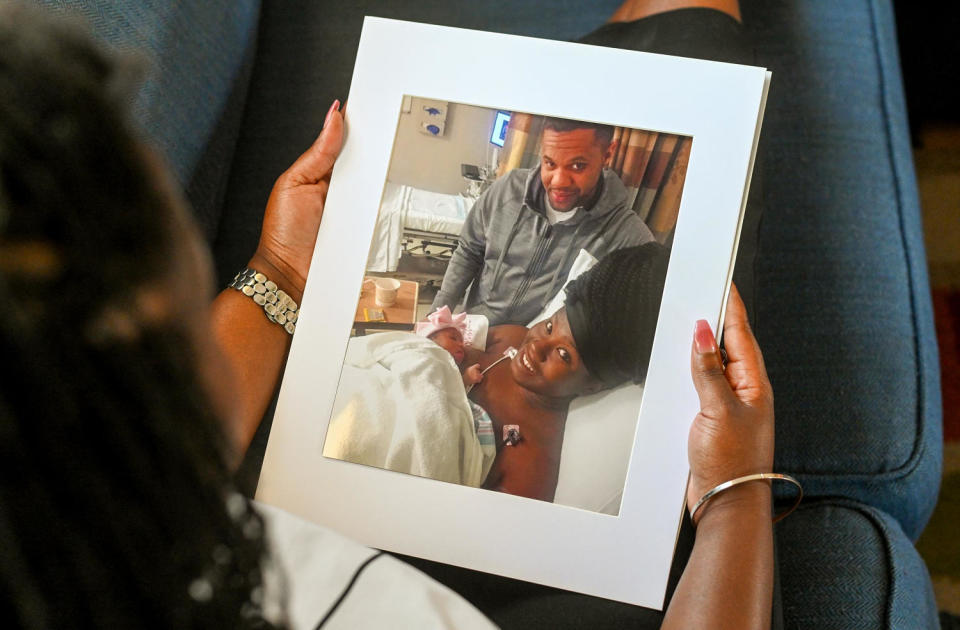 Image: Charity Watkins holds a photo of her, her newborn daughter and her husband. (Kate Medley for KFF Health News)