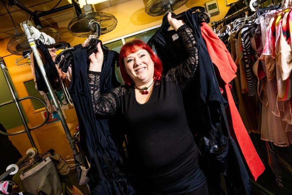 A woman in a black outfit and red hair holds up clothes on hangers.