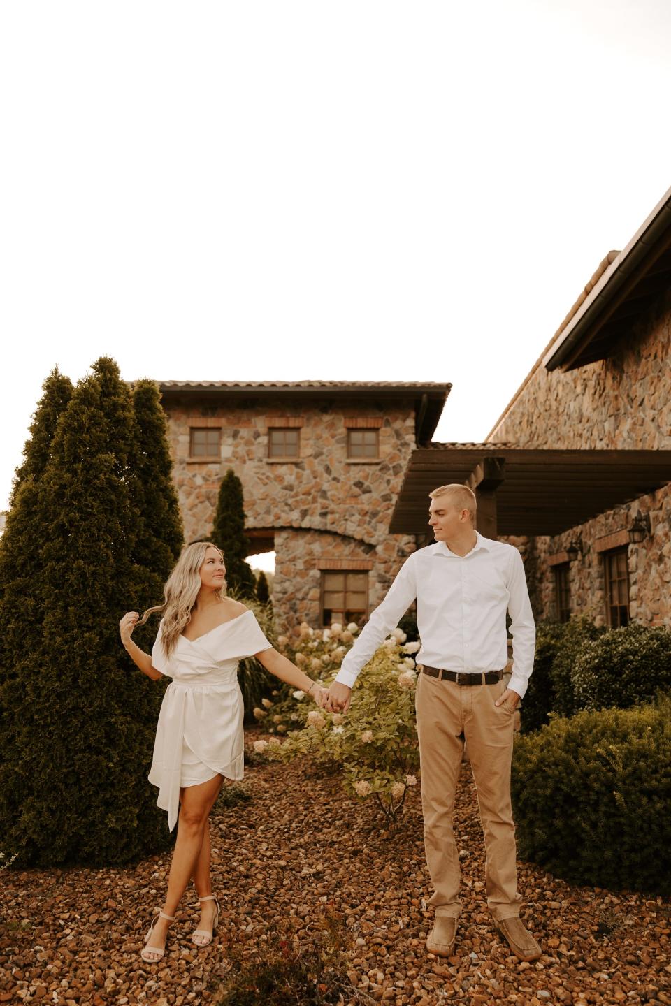 A woman in a white dress and a man in a white shirt and khakis hold hands in front of a stone building and trees.