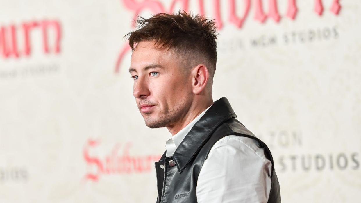 barry keoghan stands in profile with a neutral expression on his face, he wears a white collared shirt and a black leather vest, behind him is a blurry movie poster for saltburn