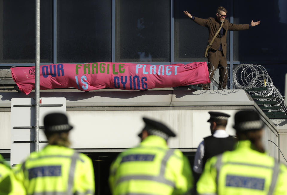 Police Officers stand guard as an Extinction Rebellion demonstrator gestures with a banner on a raised area at City Airport in London, Thursday, Oct. 10, 2019. Some hundreds of climate change activists are in London during a fourth day of world protests by the Extinction Rebellion movement to demand more urgent actions to counter global warming. (AP Photo/Matt Dunham)