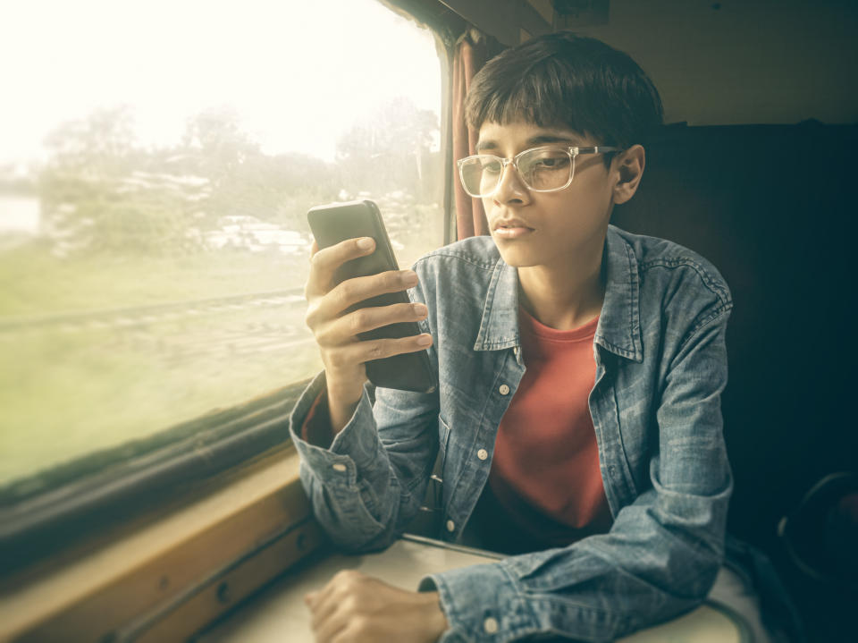 An Asian/Indian boy travels by train in the air-conditioned compartment during his summer vacation. He sits next to the window and uses a smartphone.