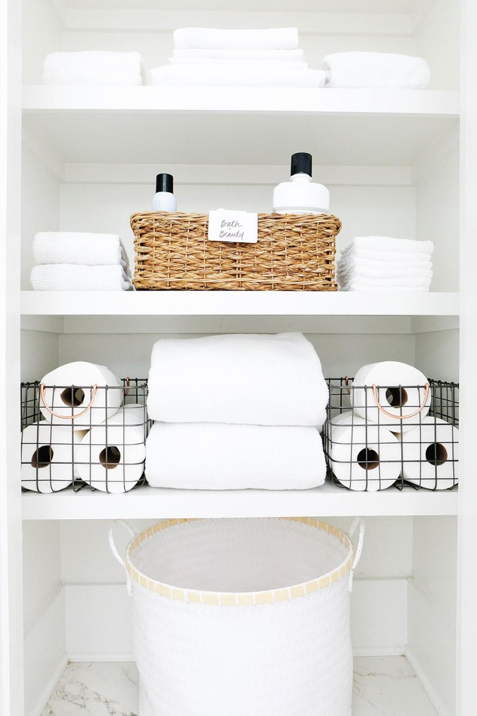 Organized Bathroom Closet Shelves with White Towels, Wicker Basket of Bath Products, Wire Basket with Toilet Paper, and White Hamper on Floor