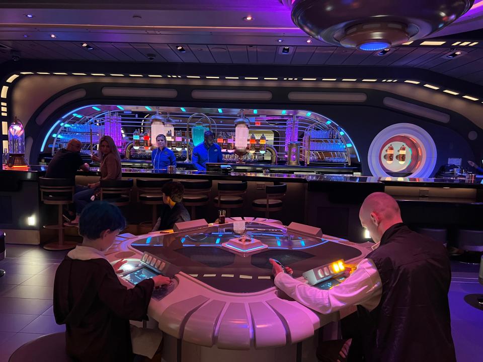 The Starcruiser lounge, with purple lighting and big tables with people pressing buttons