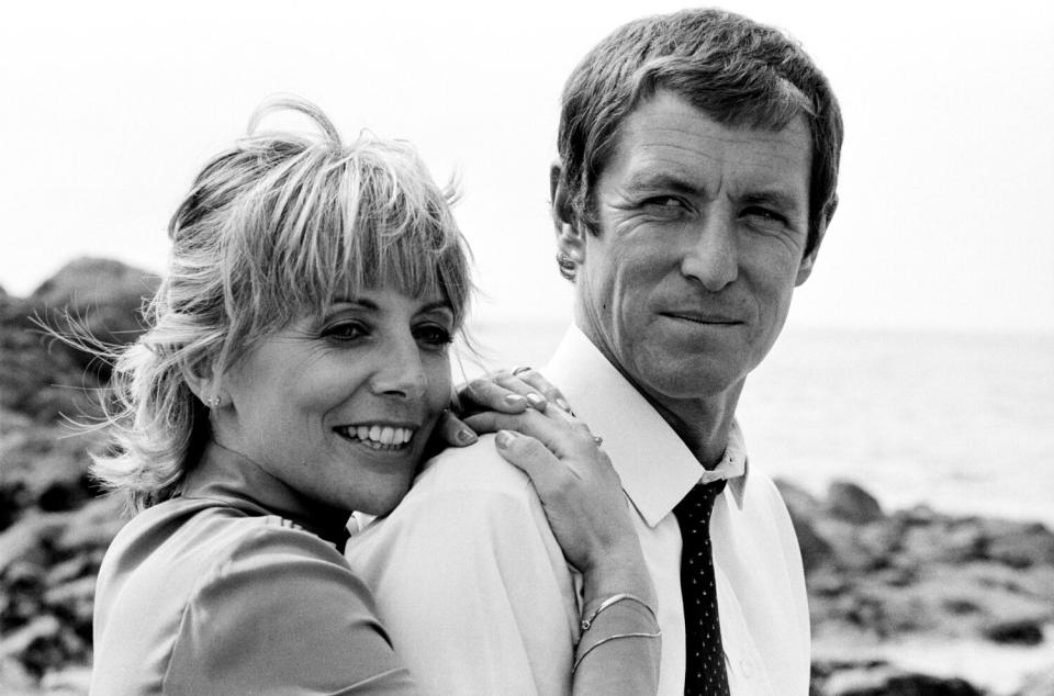 A black-and-white photo of a woman standing on the beach with her arms around a man's shoulder.