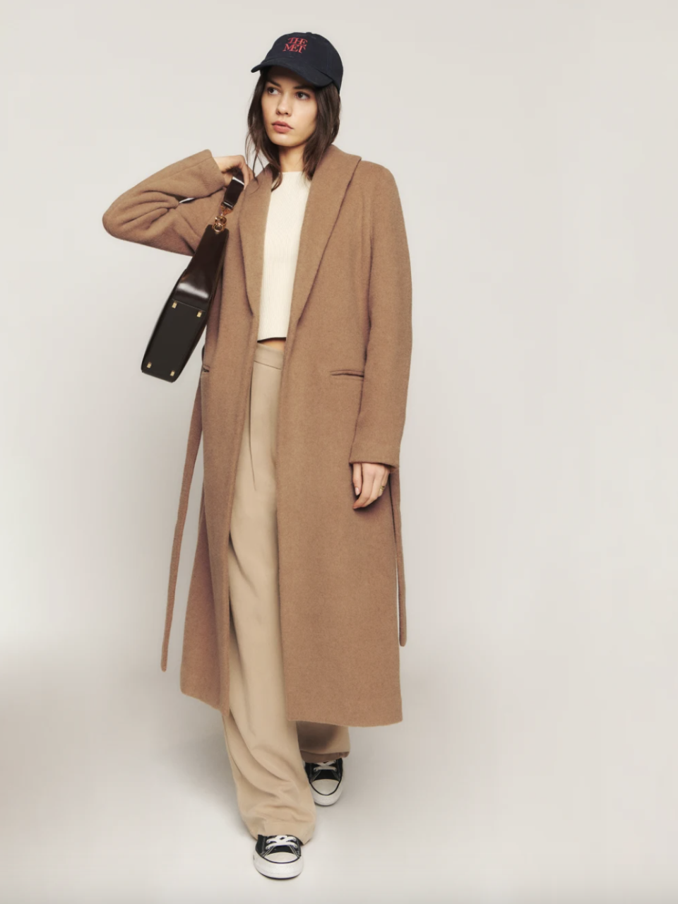 model wearing black sneakers, beige pants, white shirt and open camel reformation Downing Coat (photo via Reformation)