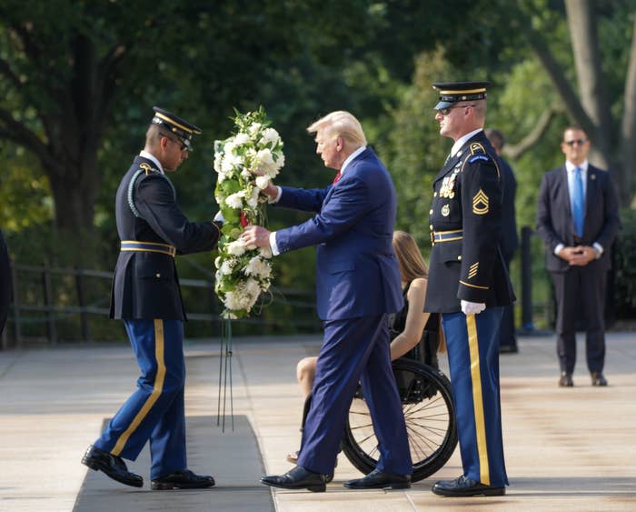 Donald Trump places a wreath with military personnel at a ceremonial event while a woman in a wheelchair and a security guard observe