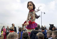 Actor Jude Law, center left, looks at Little Amal, a 3.5-meter-tall puppet of a nine-year-old Syrian girl, as it arrives at Folkestone Beach, Kent, England, Tuesday, Oct. 19, 2021 as part of the Handspring Puppet Company's 'The Walk'. The puppet started her journey in Turkey on 27 July and has travelled nearly 8,000 km across Greece, Italy, Germany, Switzerland, Belgium and France, symbolizing millions of displaced children. (Gareth Fuller/PA via AP)