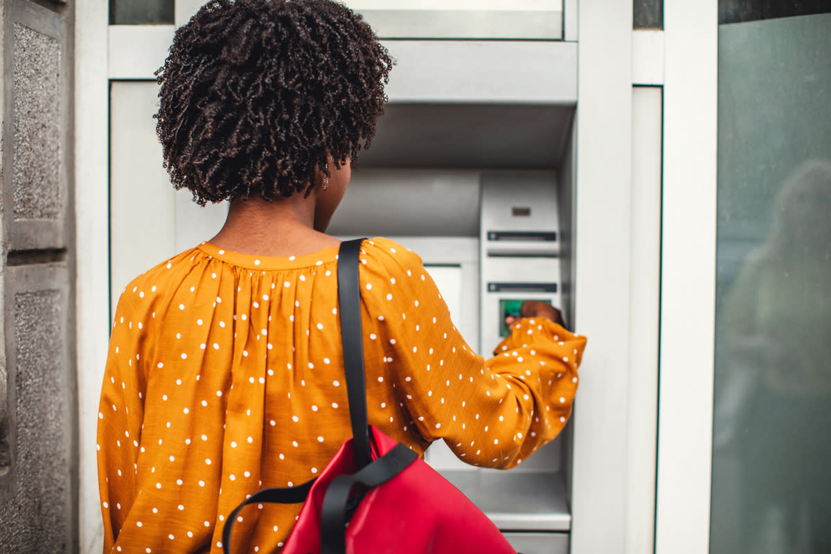 A woman with a yellow blouse and red book bag uses an ATM machine.