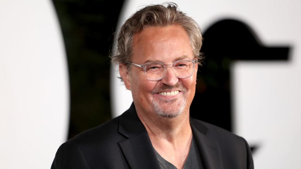 matthew perry smiles and looks right of the camera, he wears a black suit jacket over a gray t shirt and clear plastic rimmed glasses