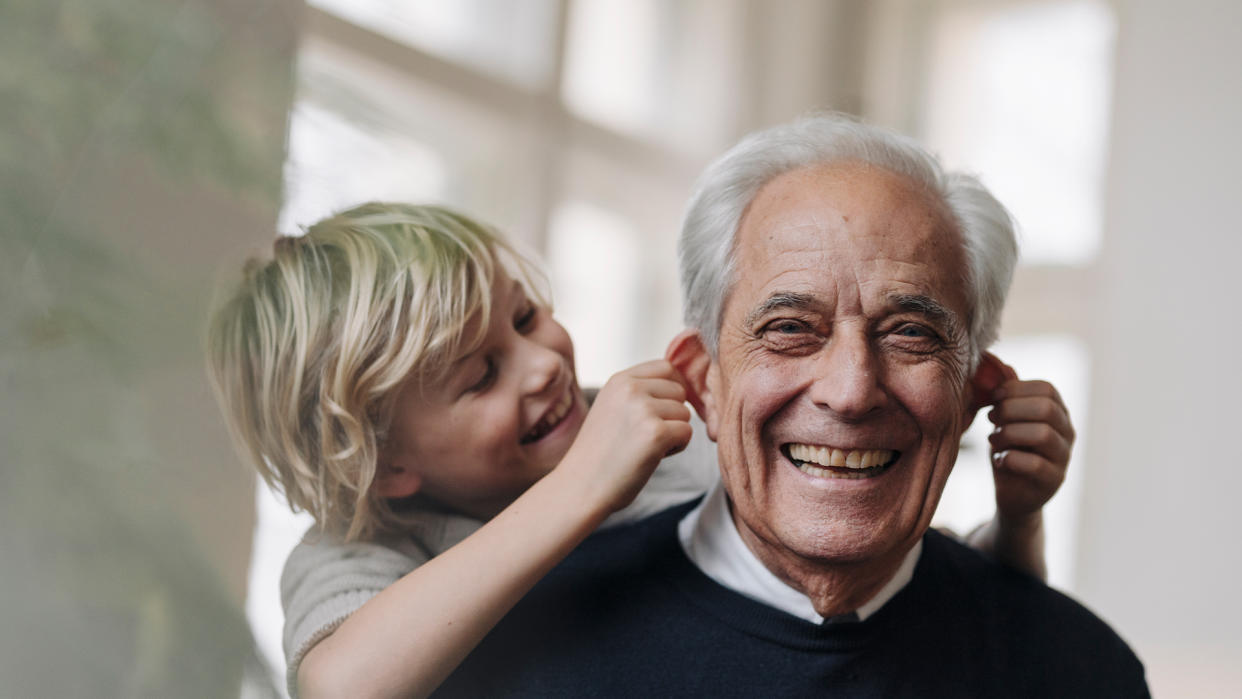  Older man gives his young grandson a piggy back ride as the child laughs and gently pulls on the grandfather's ears. 