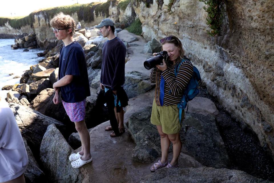 A group of people stand on rocks beside the ocean and stare at the sea. One holds a camera to her eye.