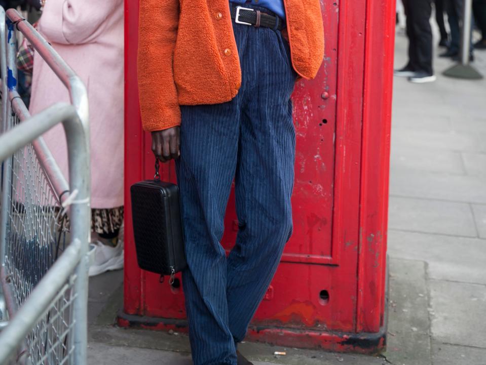 person leaning against a red wall wearing an orange cardigan, blue shirt, and blue velvet corduroys