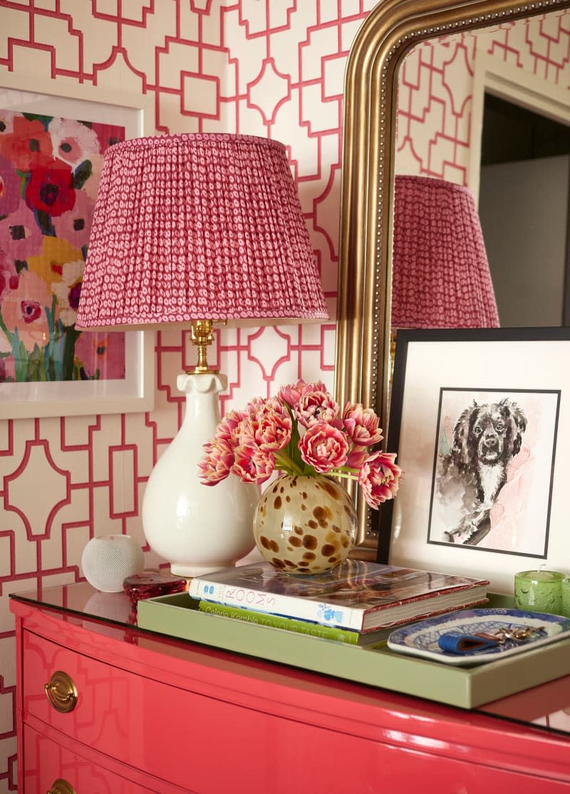 Flowers in vase and books in green tray next to lamp in front of leaning dog art and mirror on pink dresser in entryway.