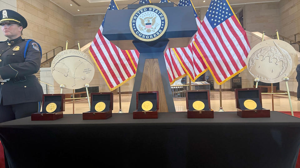 close-up photo of five gold medals lined up on a podium in front of a lectern with the presidential seal on it, with American flags in the background