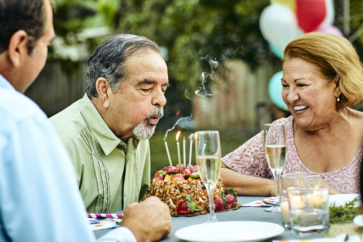 Senior woman looking at man blowing candles on birthday cake. Happy senior couple is enjoying at dining table in back yard. They are celebrating during garden party.