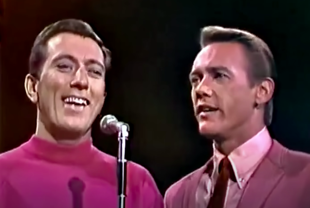 A pair of men sing into a single microphone