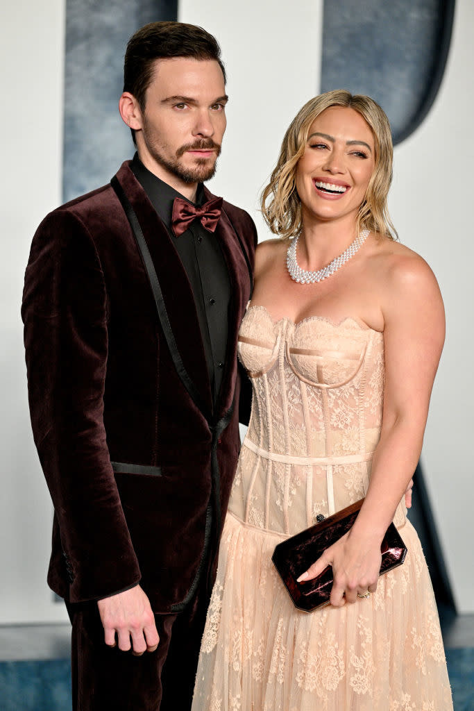 the couple on red carpet; Matthew in velvet suit, Hilary in corseted gown with pearl necklace