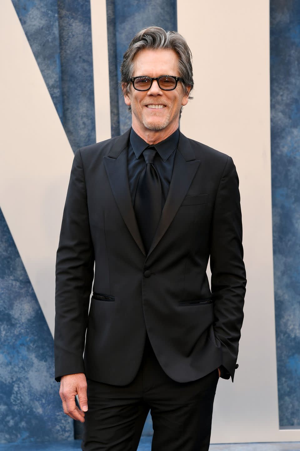 kevin bacon in a black suit and tie with tinted glasses