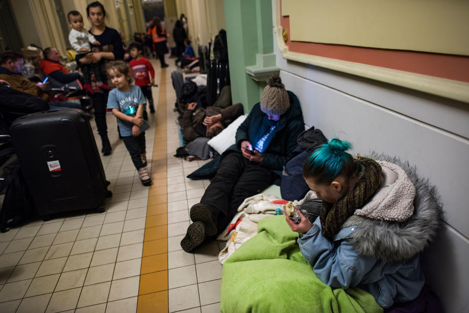 Ukrainian asylum seekers, eating or checking their phones, rest on the floor at a crowded train station.