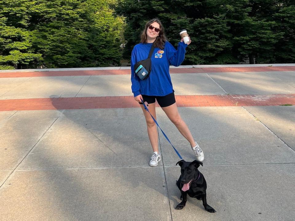 A woman standing with her dog, holding coffee and wearing a fanny pack. There are trees and a blue sky behind them.