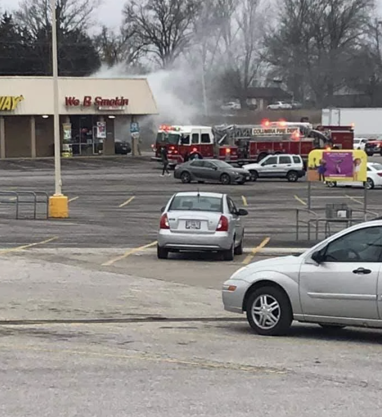 Emergency vehicles responding to a distant smoke plume in a parking lot near commercial buildings