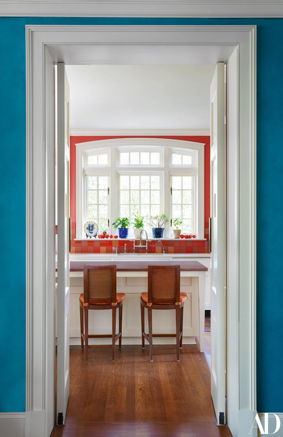 A peek into the home’s kitchen, where barstools face a window that looks out onto landscaped greenery. There had been a smaller window before, but a new one was installed in order to both bring in more light and echo the feeling of the solarium. Glass tiles in various shades of coral and red were arranged in pointillist fashion on the backsplash. “The wonderful tiler allowed me to just be insane, and we are all really pleased with it,” says Howe.