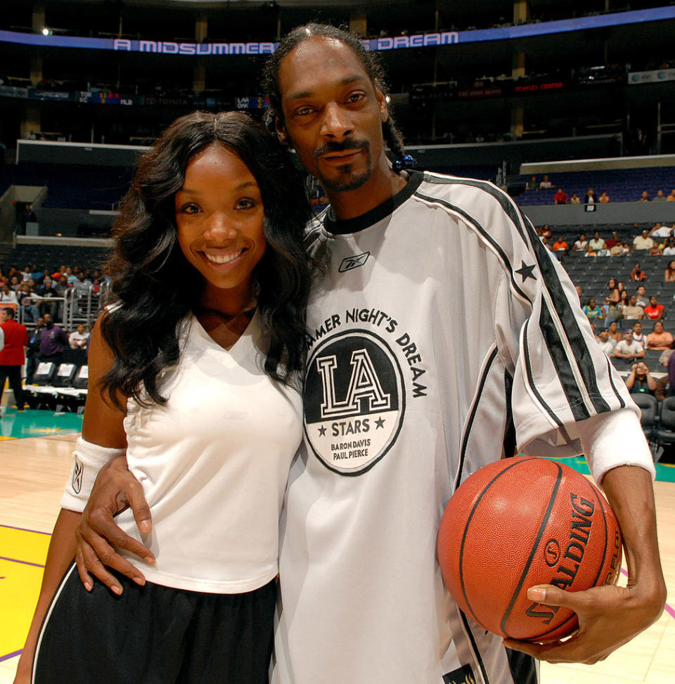 Snoop Dogg in a basketball jersey and shorts standing with a woman in a sporty white top and black pants, both smiling at a basketball event