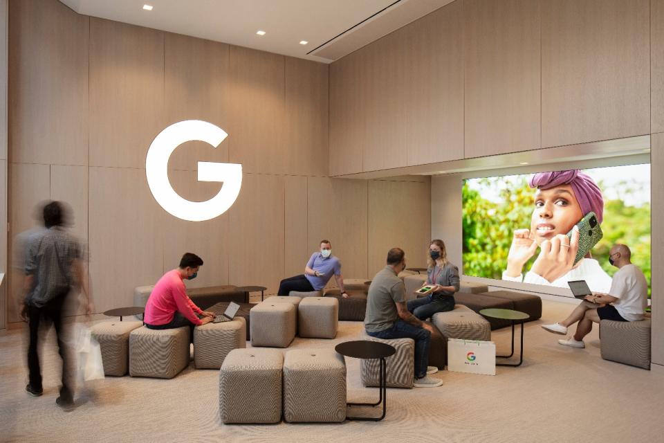 <p>Google Store Chelsea. A workshop area with several ottomans and small tables. The Google logo (letter G only) on the wall is lit in a soft white. Six people ar eseated at various spots in the room.</p>
