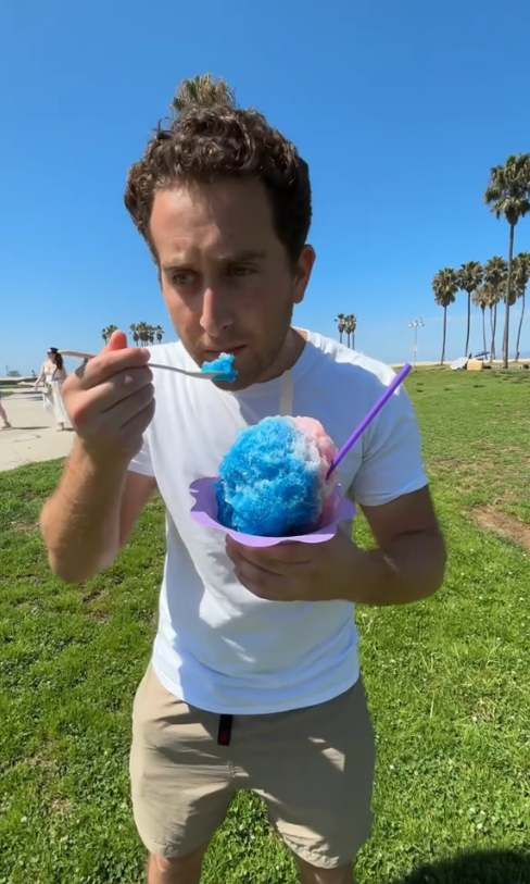 Man enjoying a large blue shaved ice treat in a park setting. Palm trees and people are visible in the background