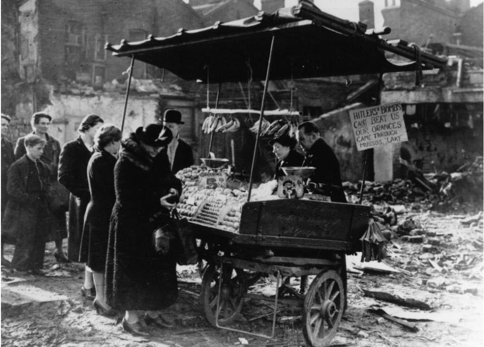 People queuing to buy oranges and bananas at a stall in bomb-wrecked London, during the Blitz, 23rd November 1940. A sign on the stall reads: 'Hitler's bombs can't beat us. Our oranges came through Musso?s lake'. Musso?s lake was service slang for the Mediterranean sea. (Photo by Keystone/Getty Images)