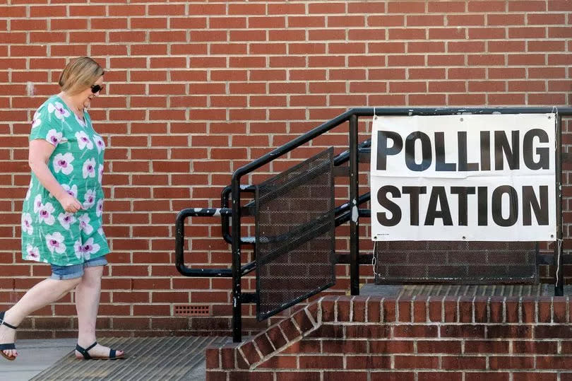 Woman in green top and denim shorts walks towards doorway with a sign outside that says 'polling station'.