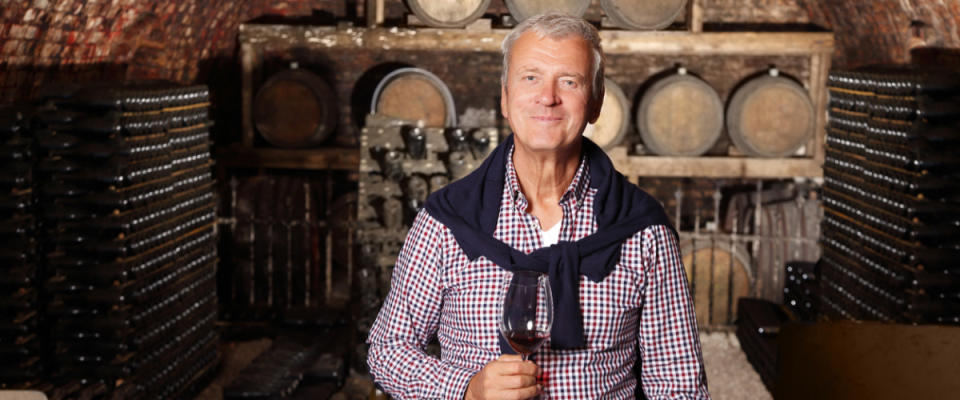 Portrait of senior standing at wine cellar while holding in his hand a glass of red wine and looking at camera