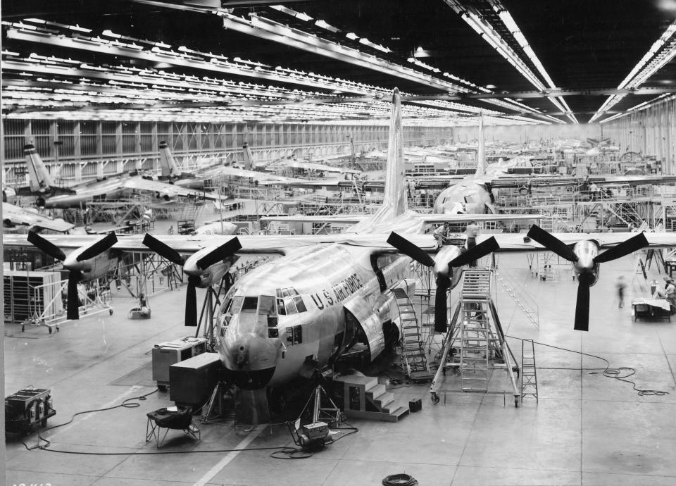 A black-and-white photo of a large cargo plane with two propellers on each side inside a large warehouse full of planes of similar sizes.