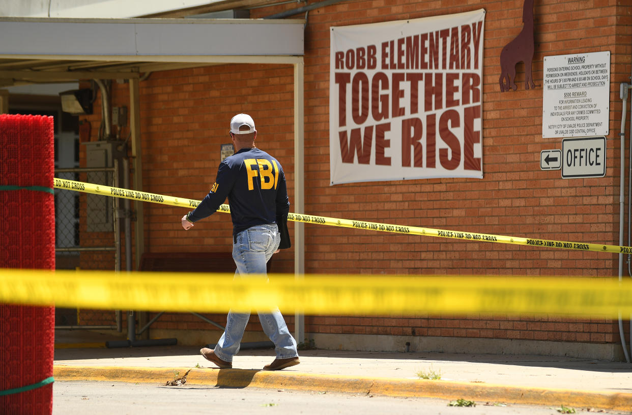 A person wearing a shirt reading FBI walks outside of a school building. A sign on the brick wall reads: Robb Elementary, Together We Rise.