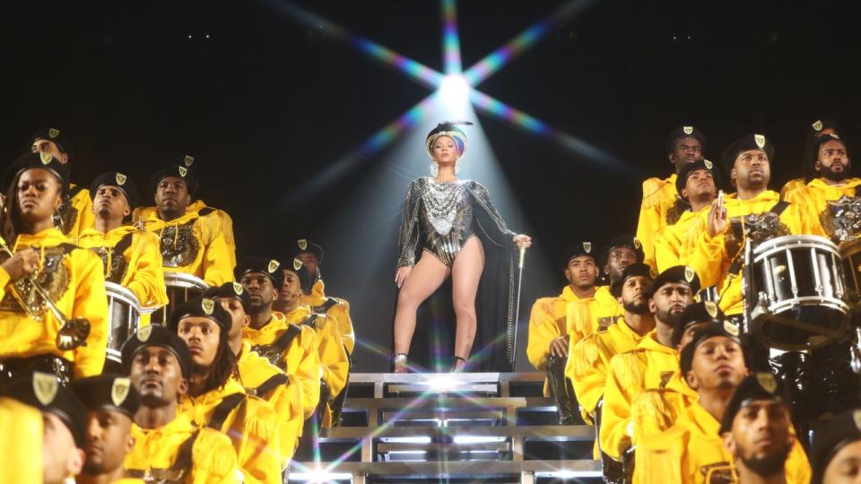 Beyonce in a queen outfit performing at Coachella. She's standing on stairs and is surrounded by dancers and a marching band wearing Yellow.