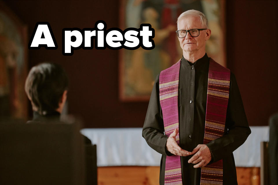 A priest wearing a clerical shirt and a patterned stole stands in a church, speaking to a person with their back to the camera