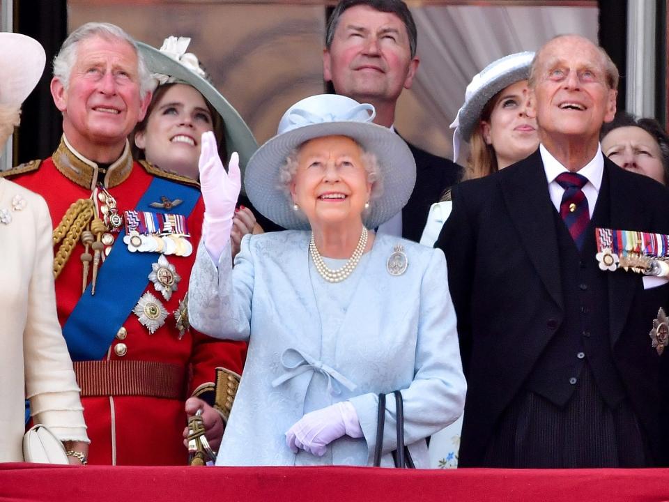 The Queen smiling and waving in a pale blue brocade overcoat with a ribbon tying it together. She has a silver oval brooch, pearl earrings and a necklace, a matching blue wide-brimmed hat with a ribbon bow on it, and white gloves.