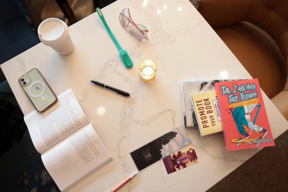 An open book, bookmarks, a cellphone, a cup of coffee, eyeglasses, a pen and stack of other books fill a table.