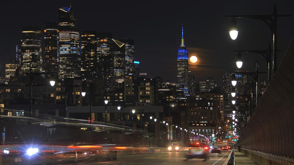     Der Vollmond geht hinter der Skyline von Downtown Manhattan und dem Empire State Building in New York auf, während der Verkehr am 25. März 2024 in Hoboken, New Jersey, das 14th Street Viaduct entlang fließt. 