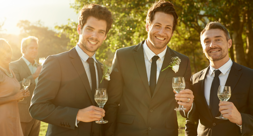 Three men from a wedding party stand holding wine glasses.