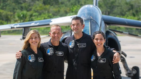 Four people in dark overalls stand in a line in front of a jet plane.