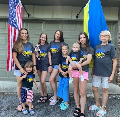 Standing between the American flag and the Ukrainian flag in front of their home, from left are, Masha and her son Artem, Mira (holding Barbie) and her mom Tania and sister Ksyusha, Olya and Nikol, and Olya’s mom Tamara.